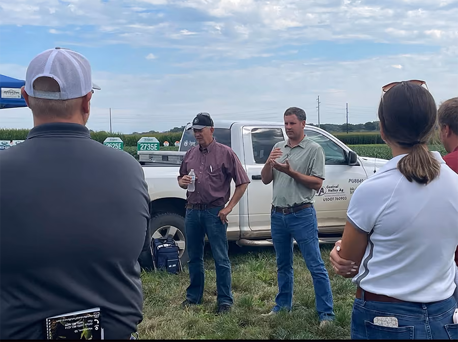 Group of people gathered outdoors on grass near a white pickup truck with company logo, two men appear to be speaking to the group on a partly cloudy day in a rural or agricultural area.