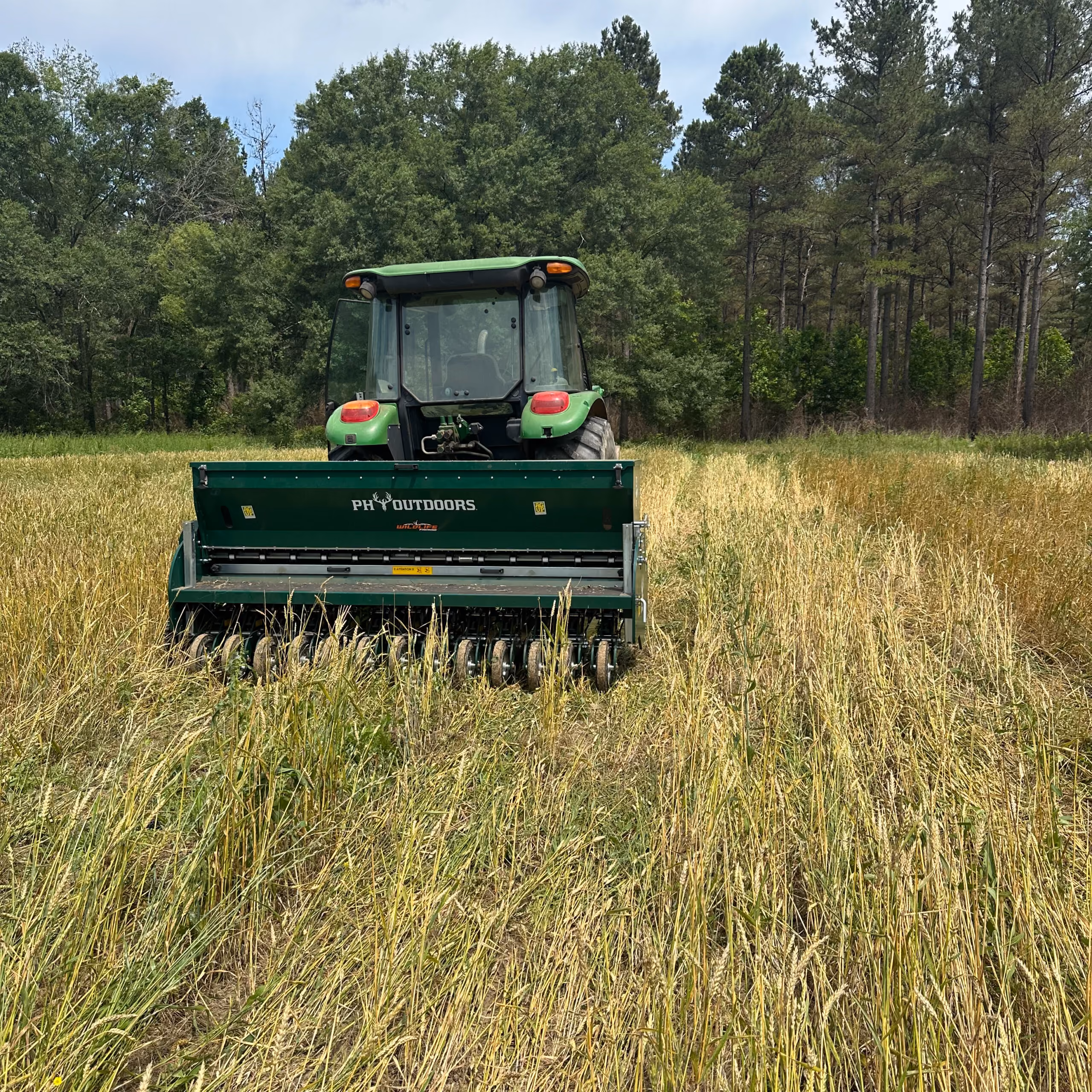 Green tractor with a PH Outdoors seeded attachment working in a field with tall grass in front of a forest with mixed green and pine trees.