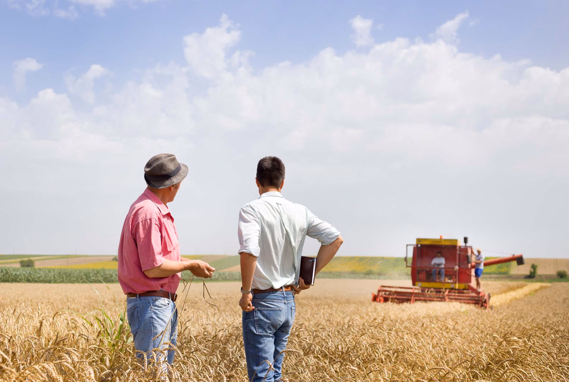 Two men standing in a golden wheat field, one wearing a pink shirt and hat, the other in a white shirt holding a notebook, observing a red combine harvester at work under a partly cloudy sky.