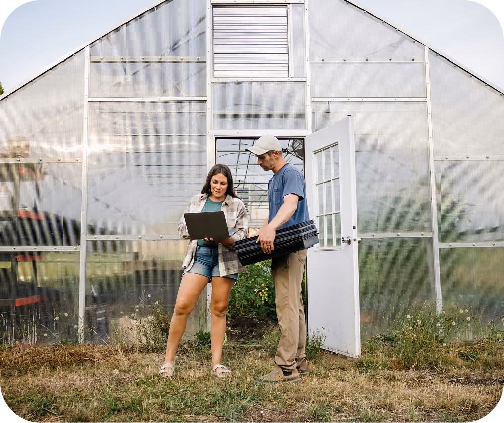 A woman and a man standing outside a greenhouse; the woman holds a laptop while the man holds black trays, both looking at the laptop screen.