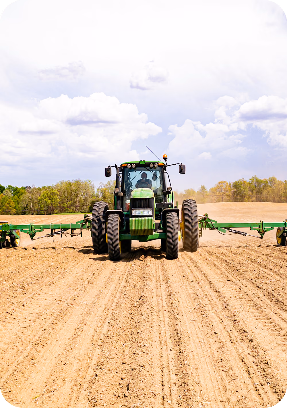 Green tractor with large tires cultivating a dusty farm field under a partly cloudy sky with a tree line in the background.