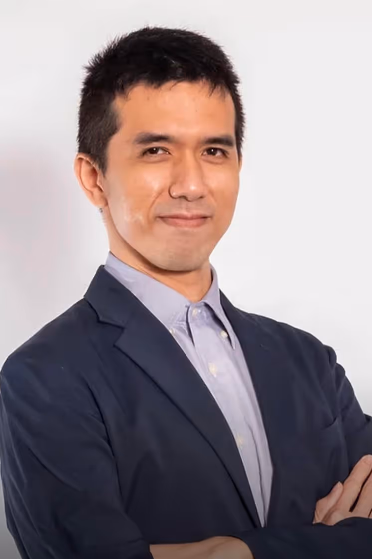 Man in a dark blazer and light shirt with arms crossed, smiling confidently against a white background.