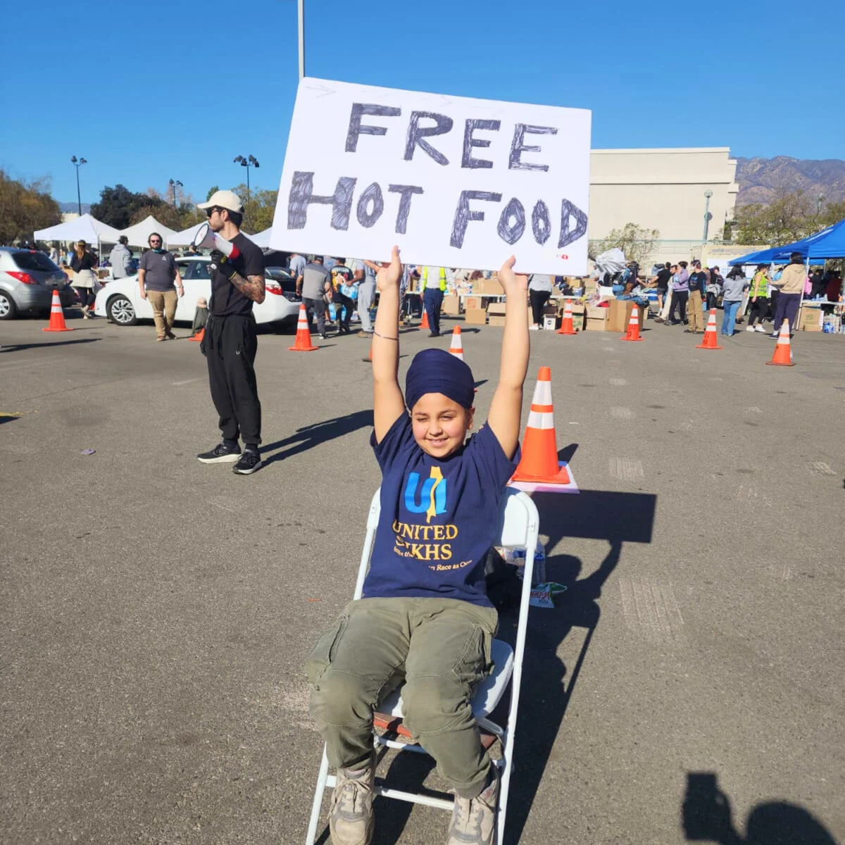A young UNITED SIKHS volunteer holding a Free Hot Food sign at an outdoor food distribution.