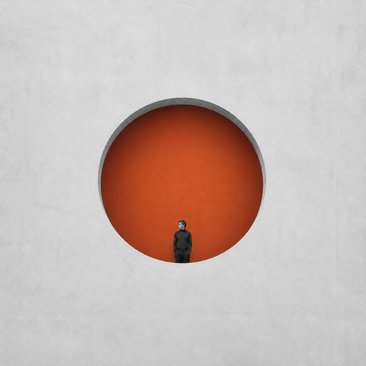 A person in black standing inside a large circular orange recess set within a smooth gray wall, looking to the left.