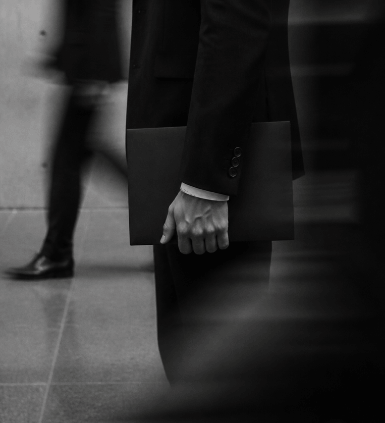 Black and white photo of a person in a suit holding a briefcase or portfolio in their hand, with blurred figures walking by in the background.