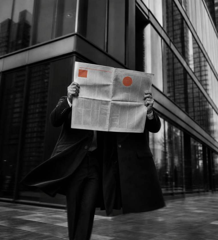Person in a business suit and coat standing outdoors in front of a modern glass building, holding a newspaper open to cover their face, with highlighted orange shapes on the paper.