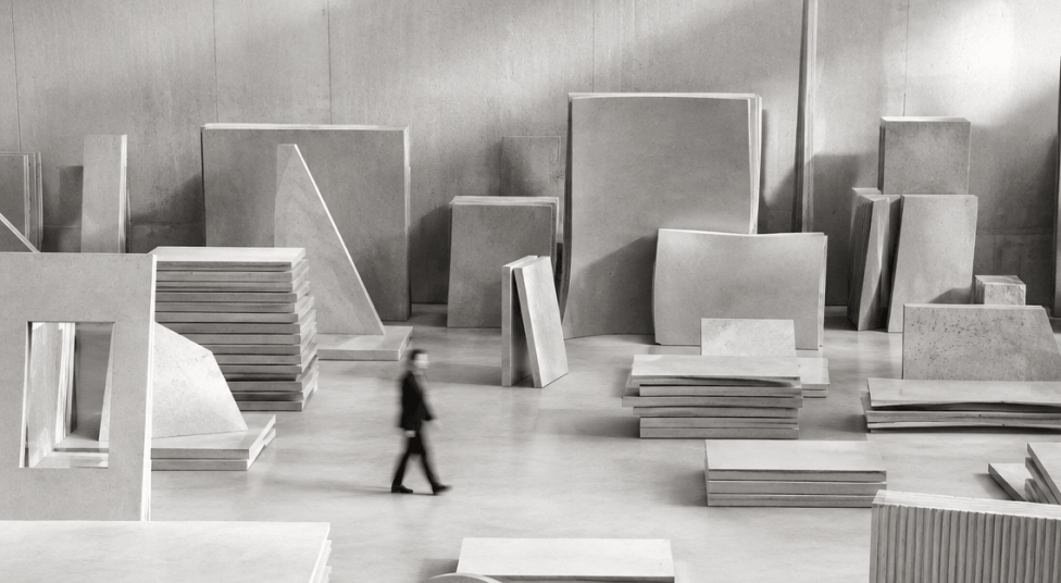 Person in black walking through large indoor space filled with many stacked and leaning rectangular and square slabs made of concrete or stone.