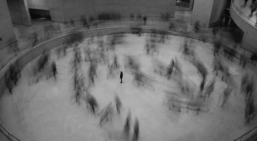 Black and white photo of a solitary person standing still in the middle of a large circular indoor space, surrounded by blurred motion of many moving people.