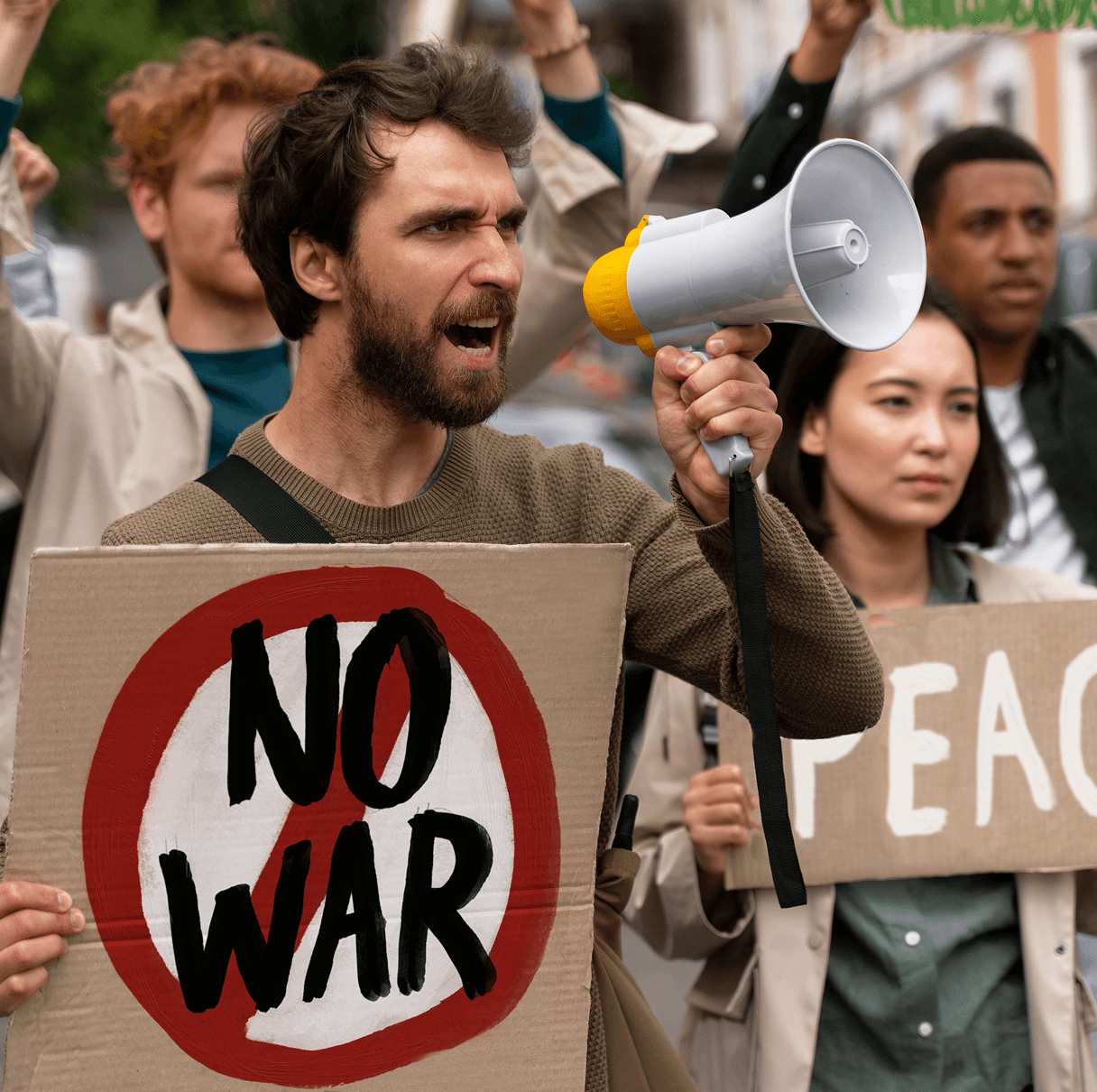 Man holding a megaphone and a sign that reads 'NO WAR' at a protest, with other demonstrators holding signs including one that says 'PEACE'.
