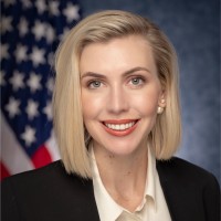 Ashley Zumwalt-Forbes in business attire smiling with American flag in the background.