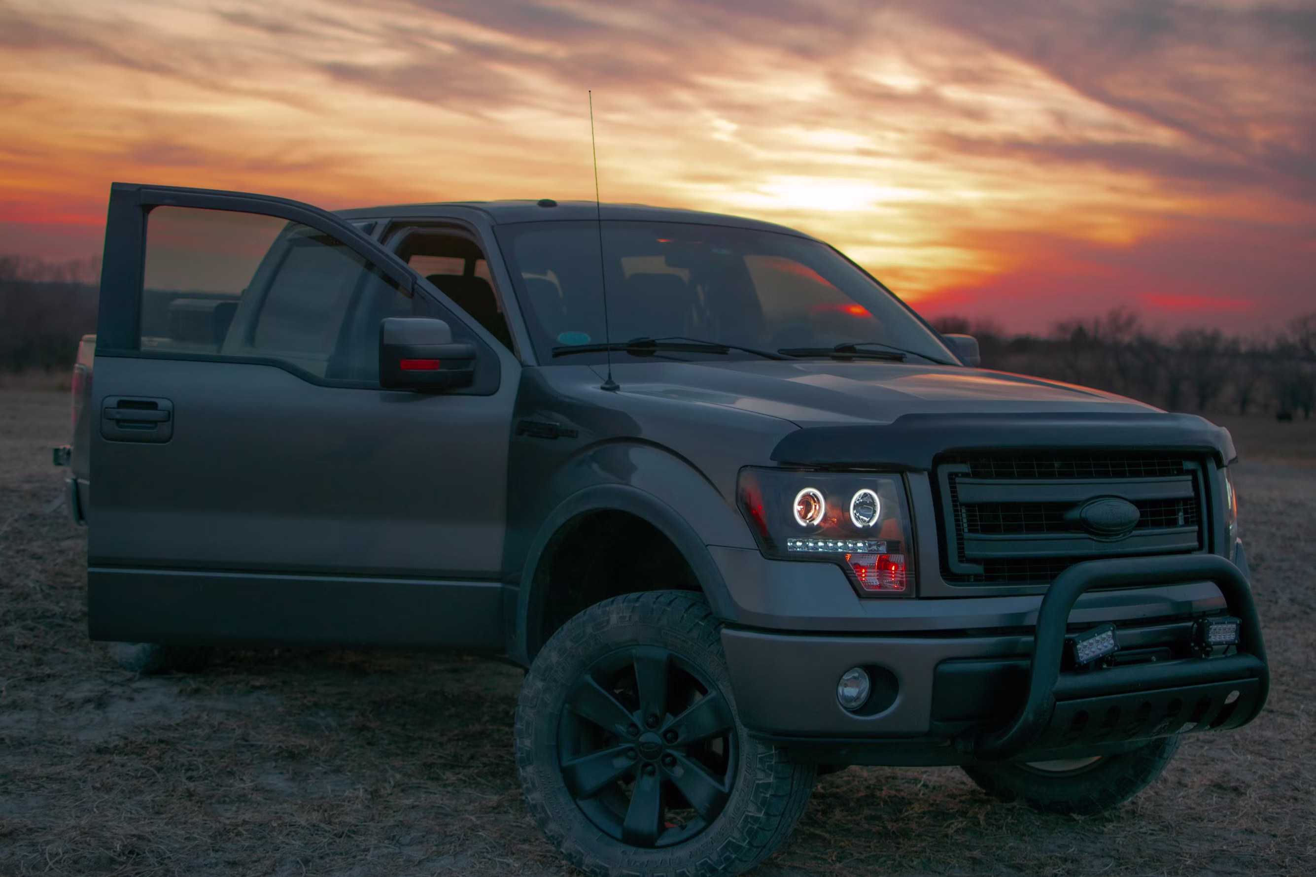 Black pickup truck with front door open on dirt ground at sunset with cloudy sky.