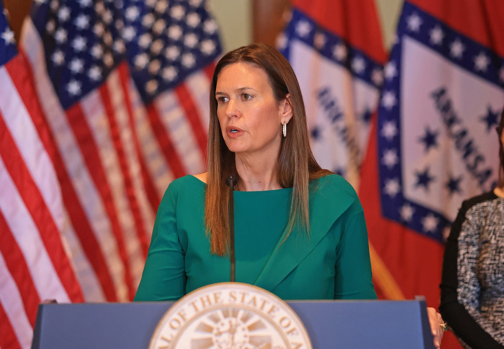 Sarah Huckabee Sanders in green speaking at a podium with U.S. and Arkansas state flags behind her.