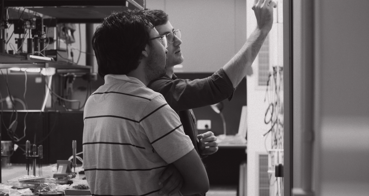 Two men facing a whiteboard, one is writing on it