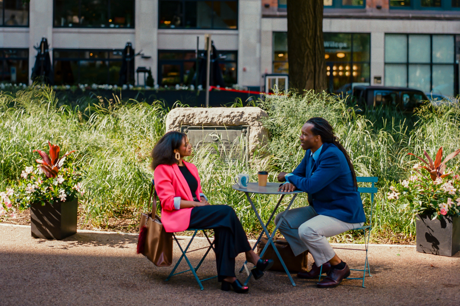 A man in a blue jacket and a woman in a bright pink jacket sit at a bistro table in a park.