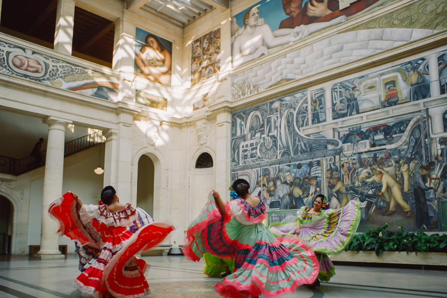 Dancers wearing colorful dresses spin in unison, in a two-story atrium covered in murals.