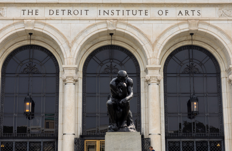 Photo of "The Detroit Institute of Arts" engraved into the building above three arched windows. A statue is in foreground.