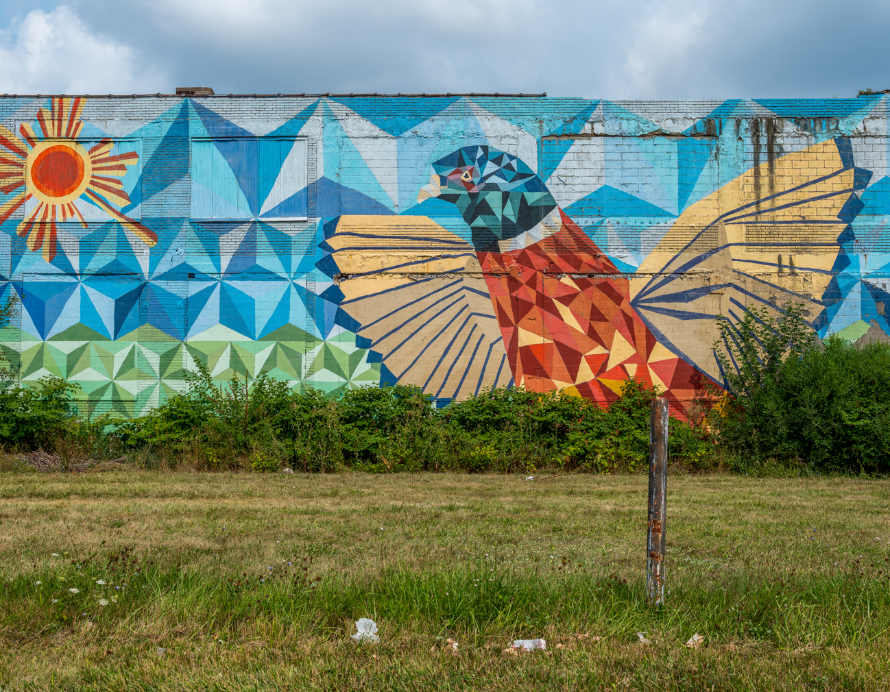 A photo of a brick wall mural in a fractal art style featuring a large orange and blue bird facing an image of the sun.