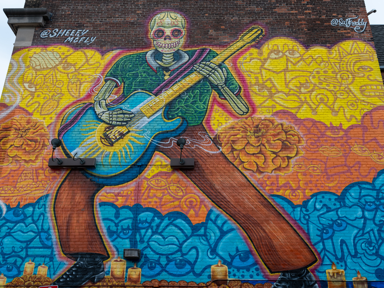 Photo of a brick wall painted with a mural of a skeleton playing a guitar on top of a yellow, orange, and blue sectioned background.