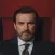 Portrait of a man with a beard and mustache wearing a suit and tie against a dark red background.