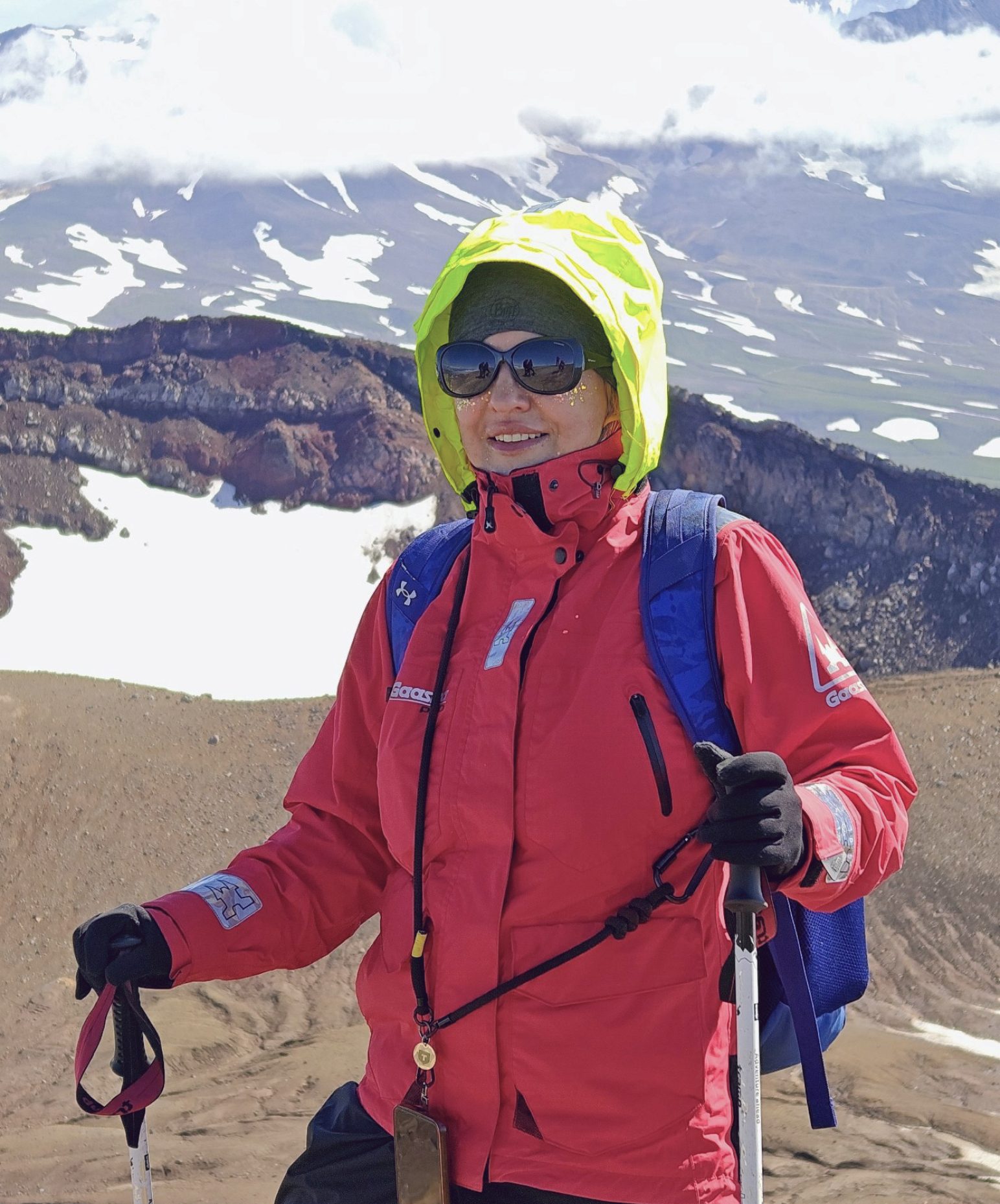 Elena Vysotskaia on a volcanic ridge in a red jacket, with snowfields and cloud behind her