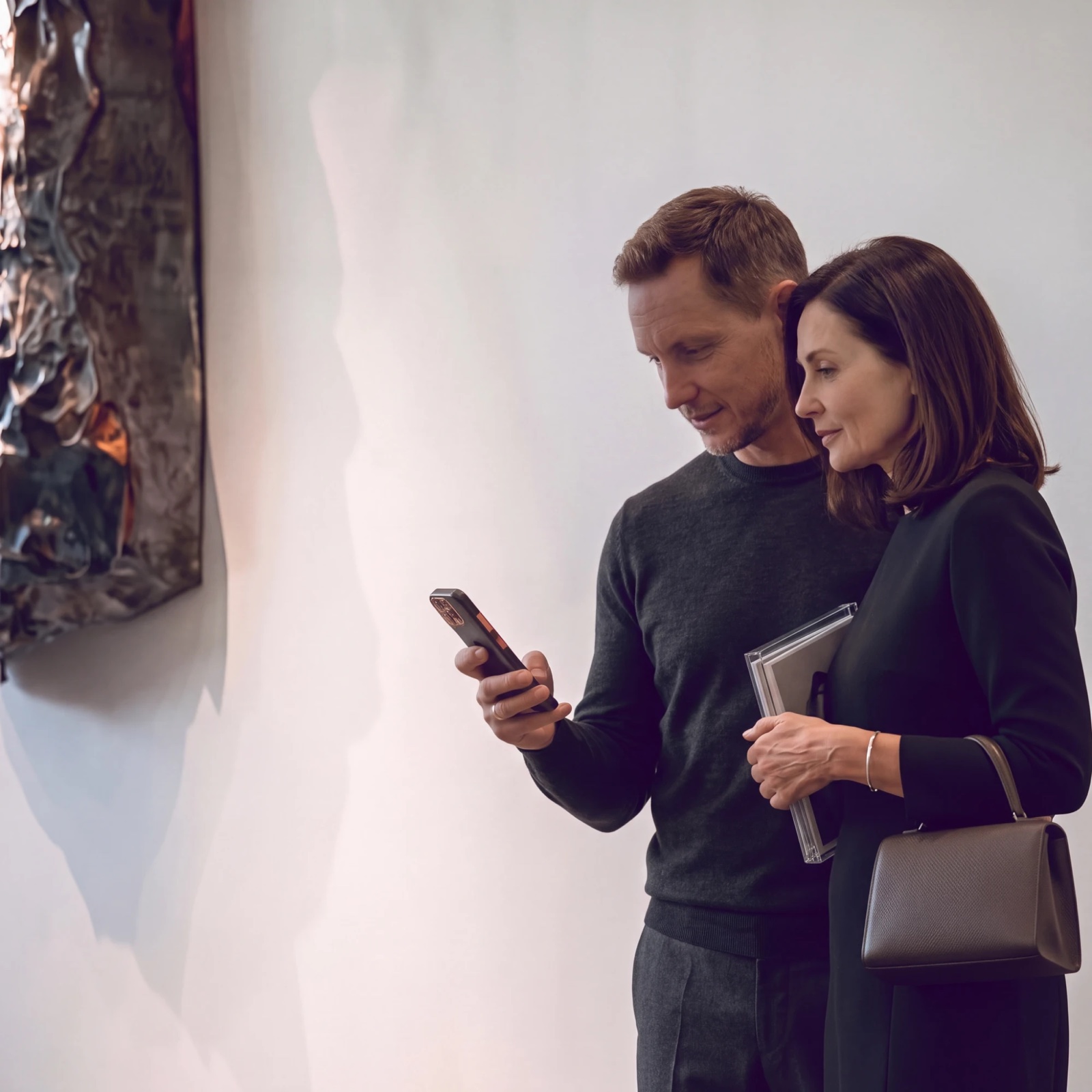 A man and a woman looking at a phone together beside a crumpled metal wall sculpture