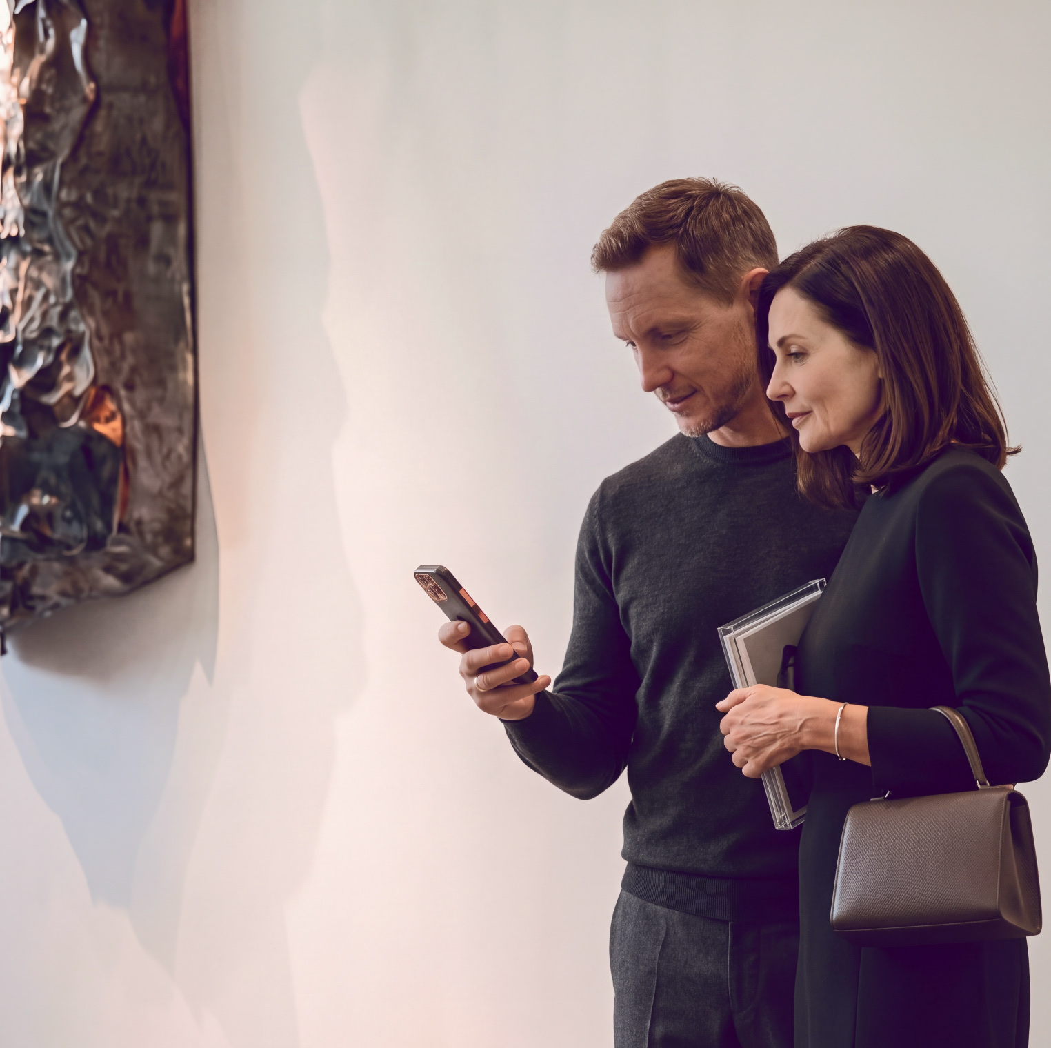 A man and a woman looking at a phone together beside a crumpled metal wall sculpture