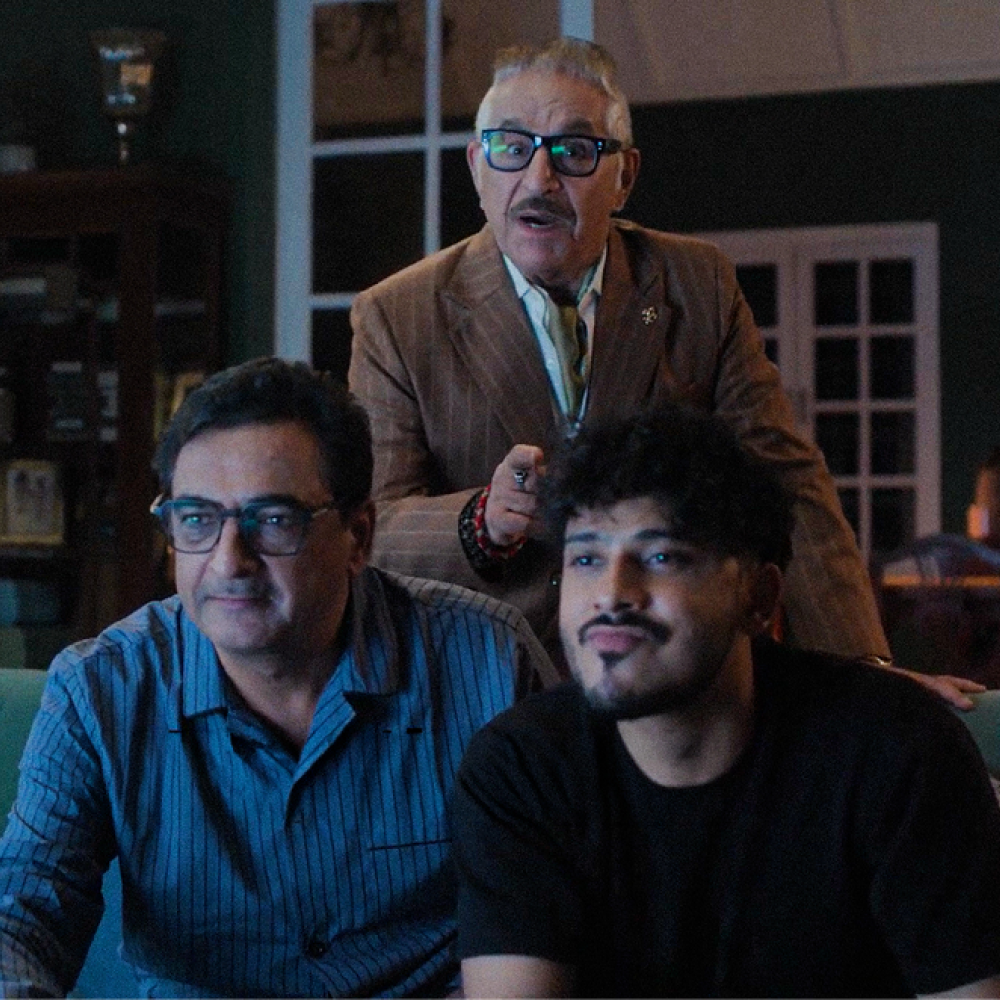 Three men indoors with an older man in a brown suit and glasses standing behind two younger men sitting on a couch, one wearing a blue striped shirt and the other a black shirt.