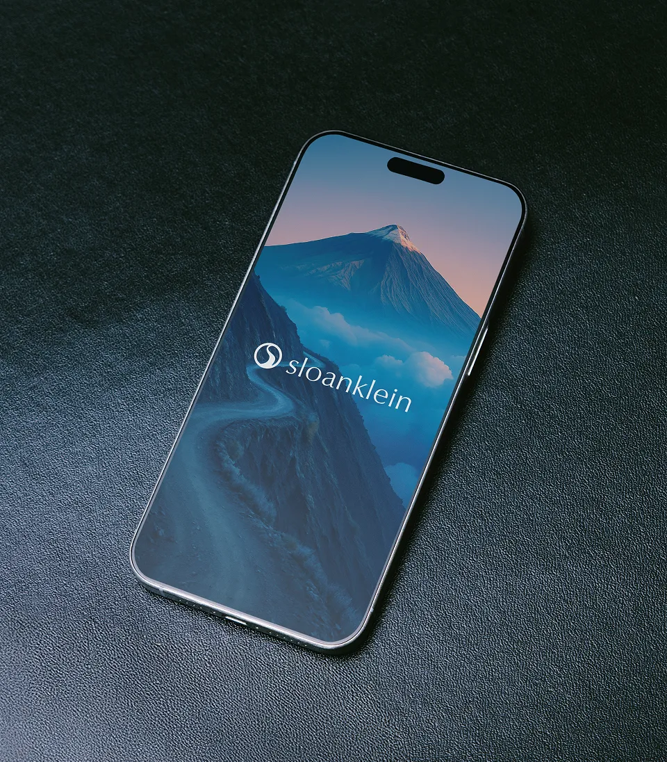 Smartphone on dark surface displaying a mountain landscape with a winding road and the Sloan Klein logo.