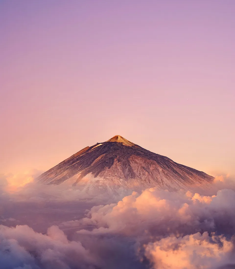 Mountain peak above purple and orange clouds at sunrise or sunset with a pink sky background.