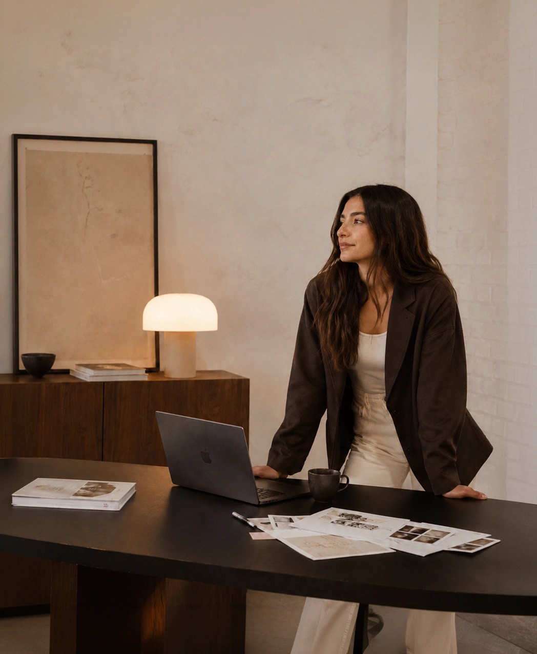 Woman with long brown hair leaning on black table with laptop, papers, cup, and warm lamp in background.