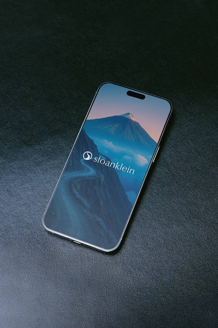 Smartphone on black surface showing mountain landscape with sloan klein logo on screen.