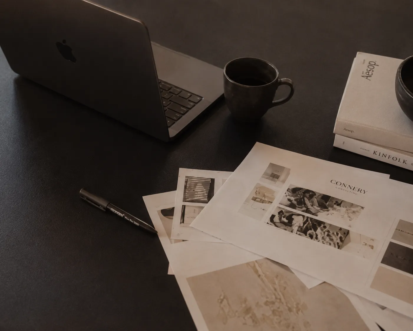 Laptop, coffee cup, pen, and printed papers with design layouts on a dark desk.