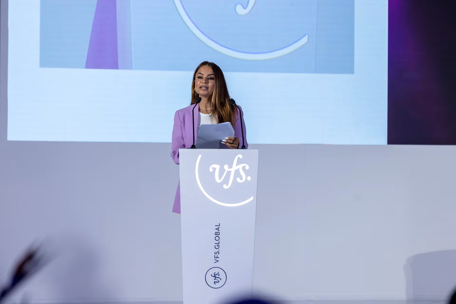 Woman speaking at a VFS Global event, standing at a branded podium with a presentation screen behind her.