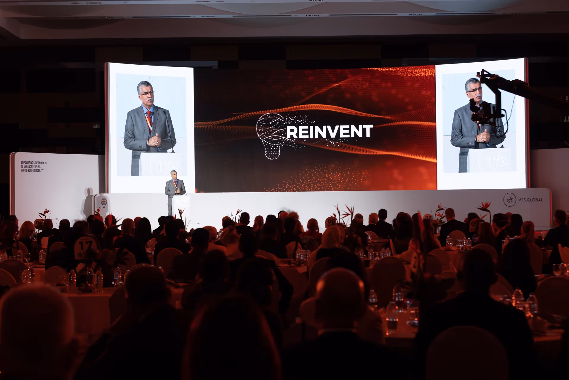 Speaker presenting on stage at a VFS Global event, with large screens displaying the “Reinvent” theme and a seated audience in the foreground.