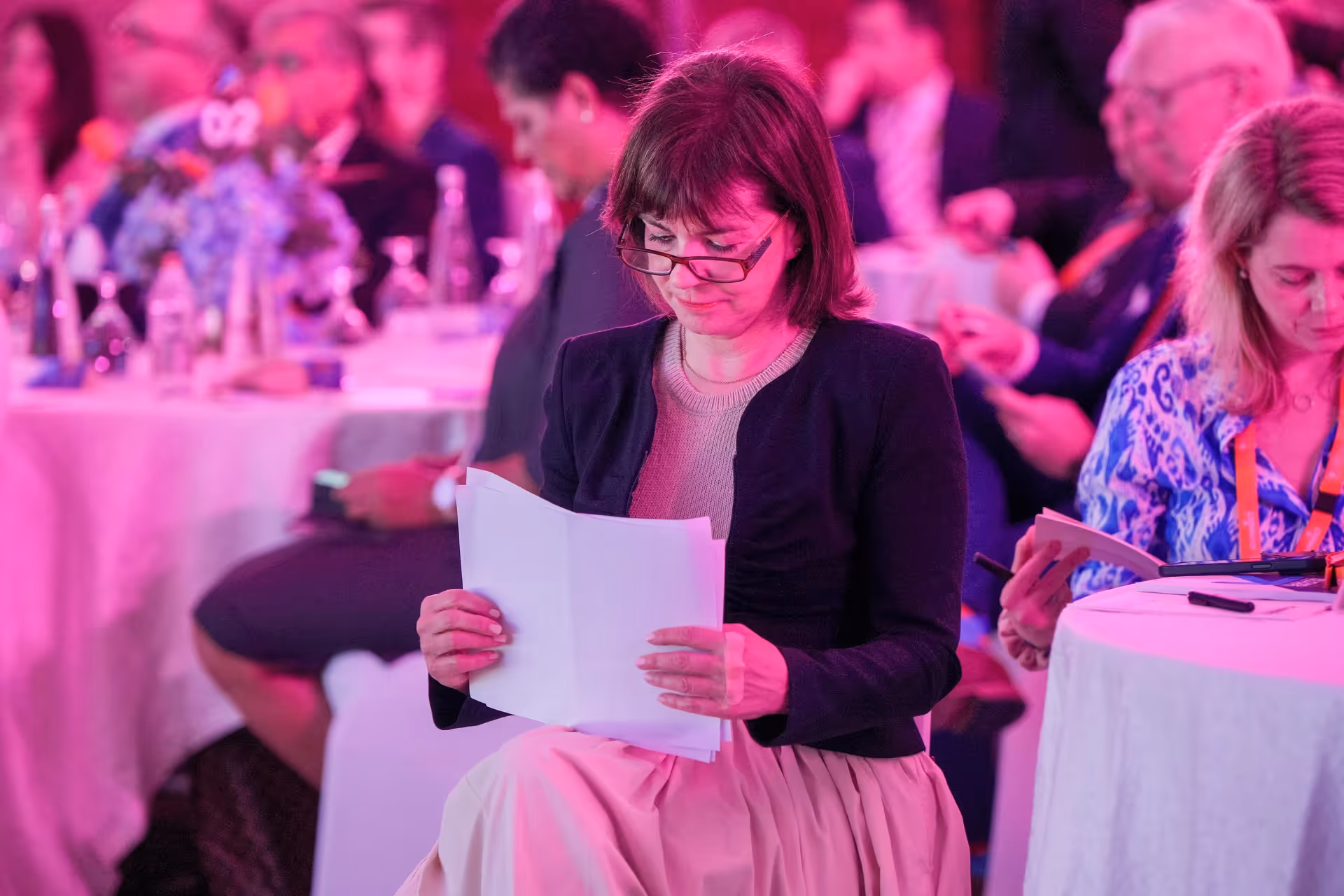 Conference attendee reviewing printed materials while seated at a networking event, with other attendees gathered at tables under pink lighting.
