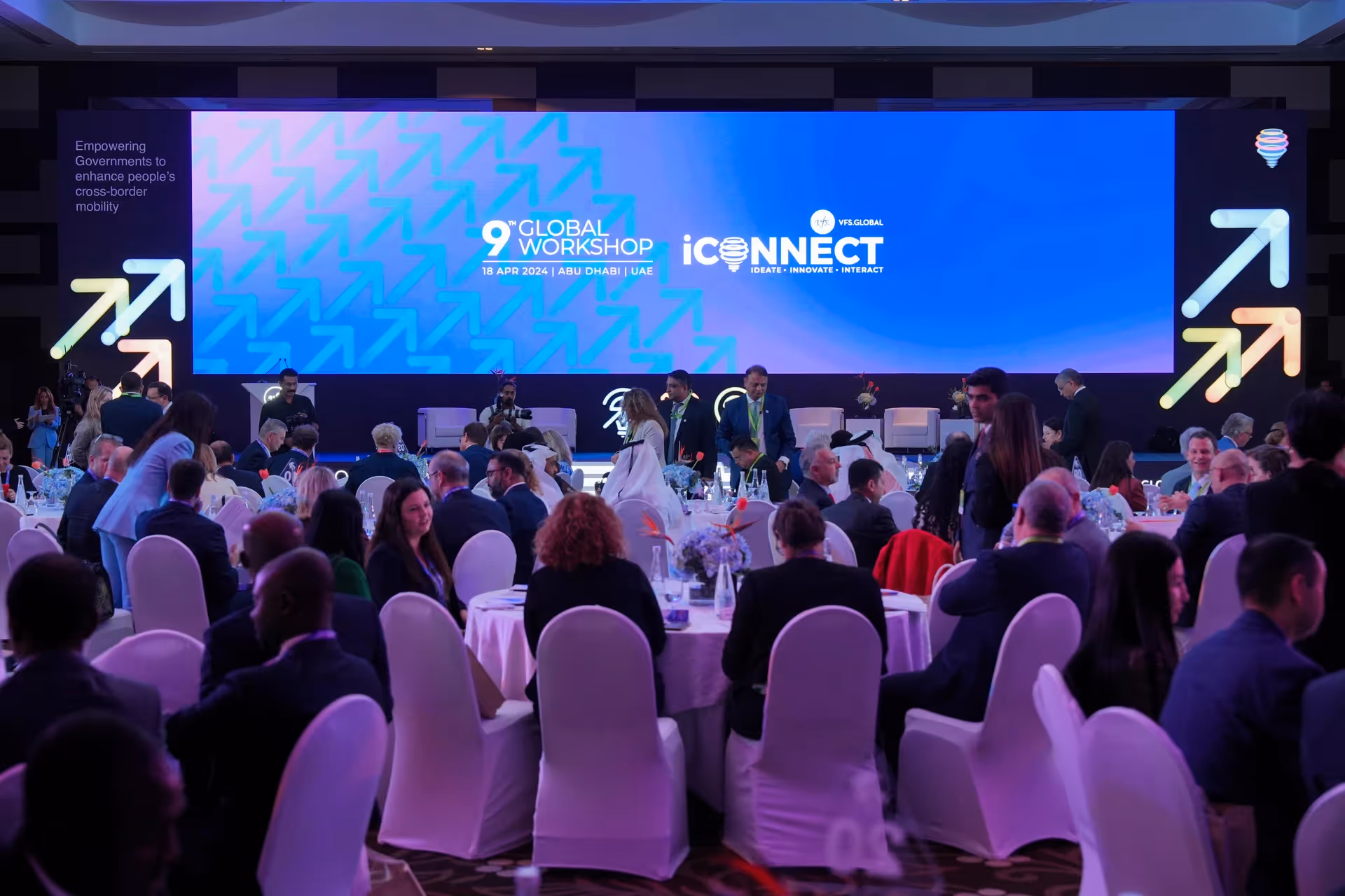 Audience seated in an event venue watching a speaker present on stage beneath large digital screens.