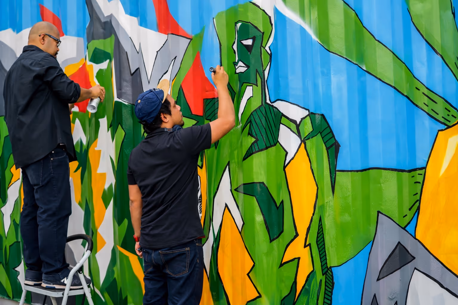 Two artists paint a colorful comic-style mural of a green superhero on a blue wall; one stands on a ladder while the other paints details.