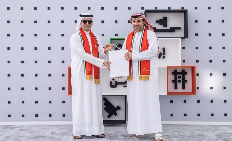 Two men in traditional Emirati attire presenting a certificate at a UAE cultural event, wearing red-and-white scarves.