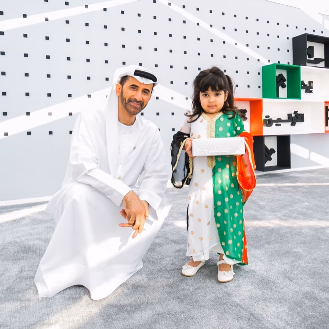Emirati man in traditional attire posing with a young girl in a green and white traditional dress, who is holding a certificate at a cultural event.