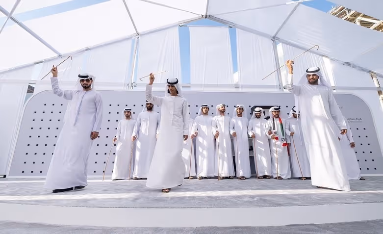 Traditional Emirati performers in white kanduras participating in a cultural dance with canes at an outdoor event.