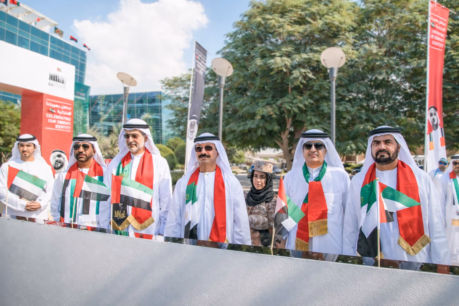 Group of people gathered around a UAE flag during a flag-raising ceremony outside a modern building at night.