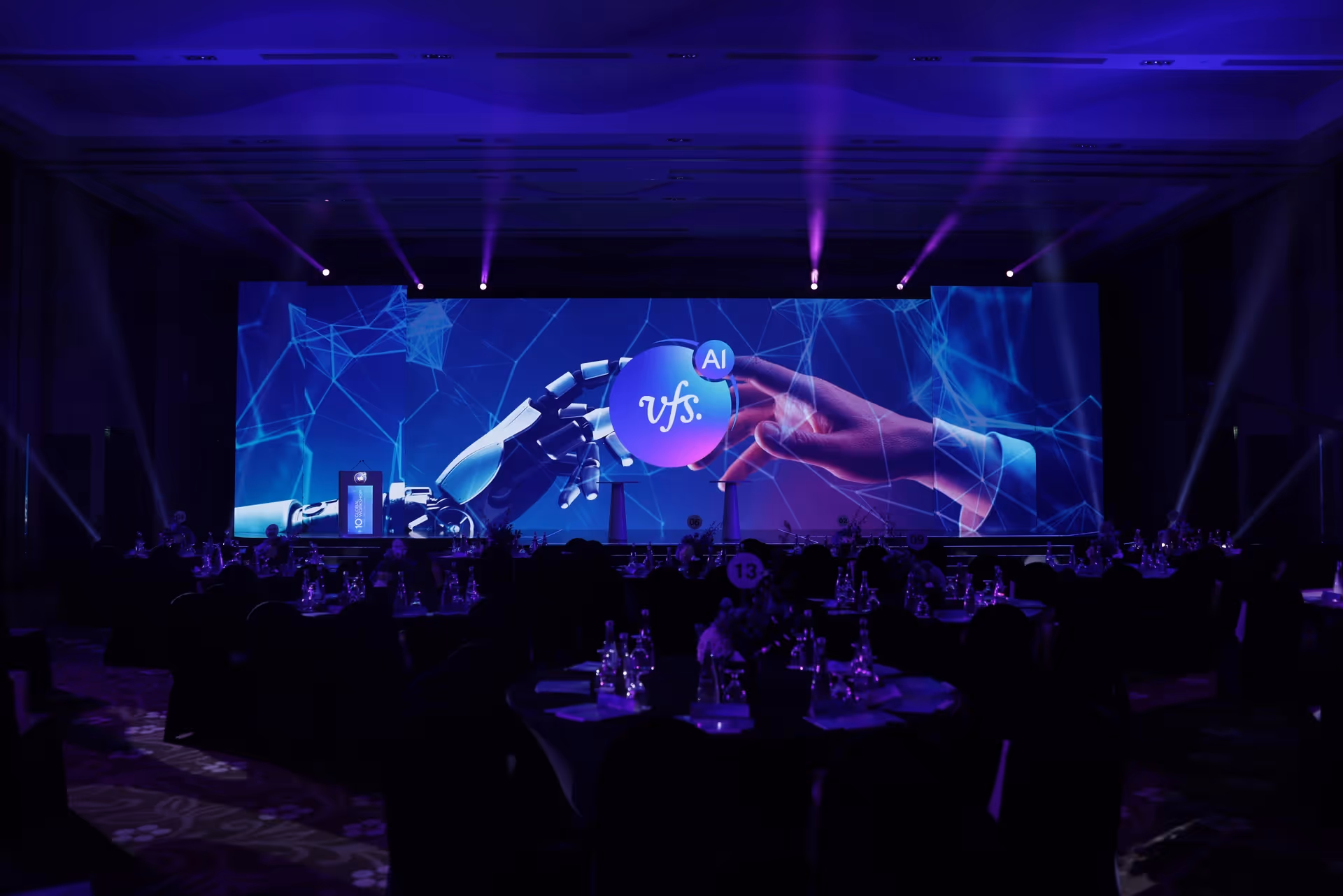 Audience seated at a technology event with a large digital stage display featuring a robotic hand, human hand, and AI-themed visuals.