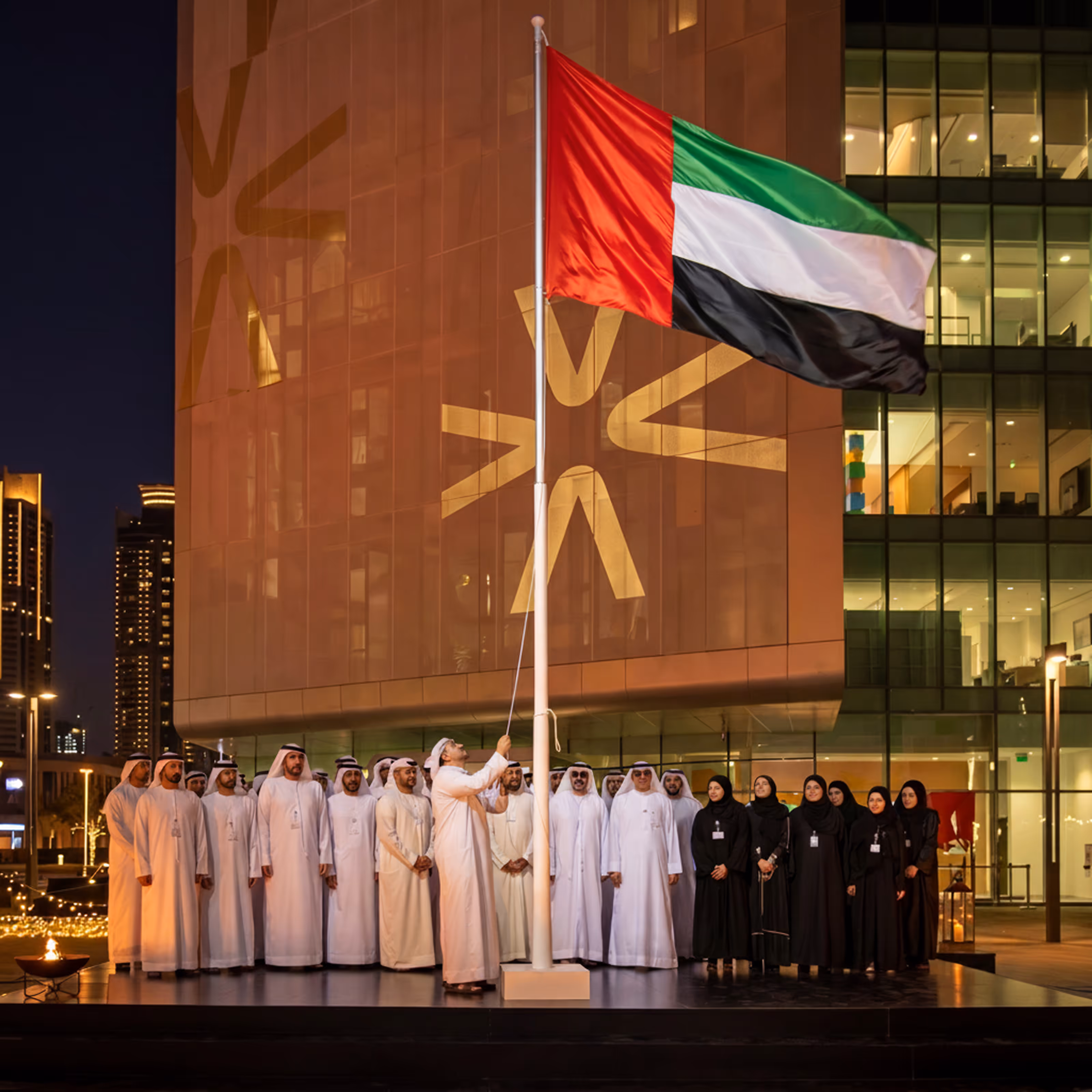 Group of people gathered for a UAE flag-raising ceremony outside a modern building at night.