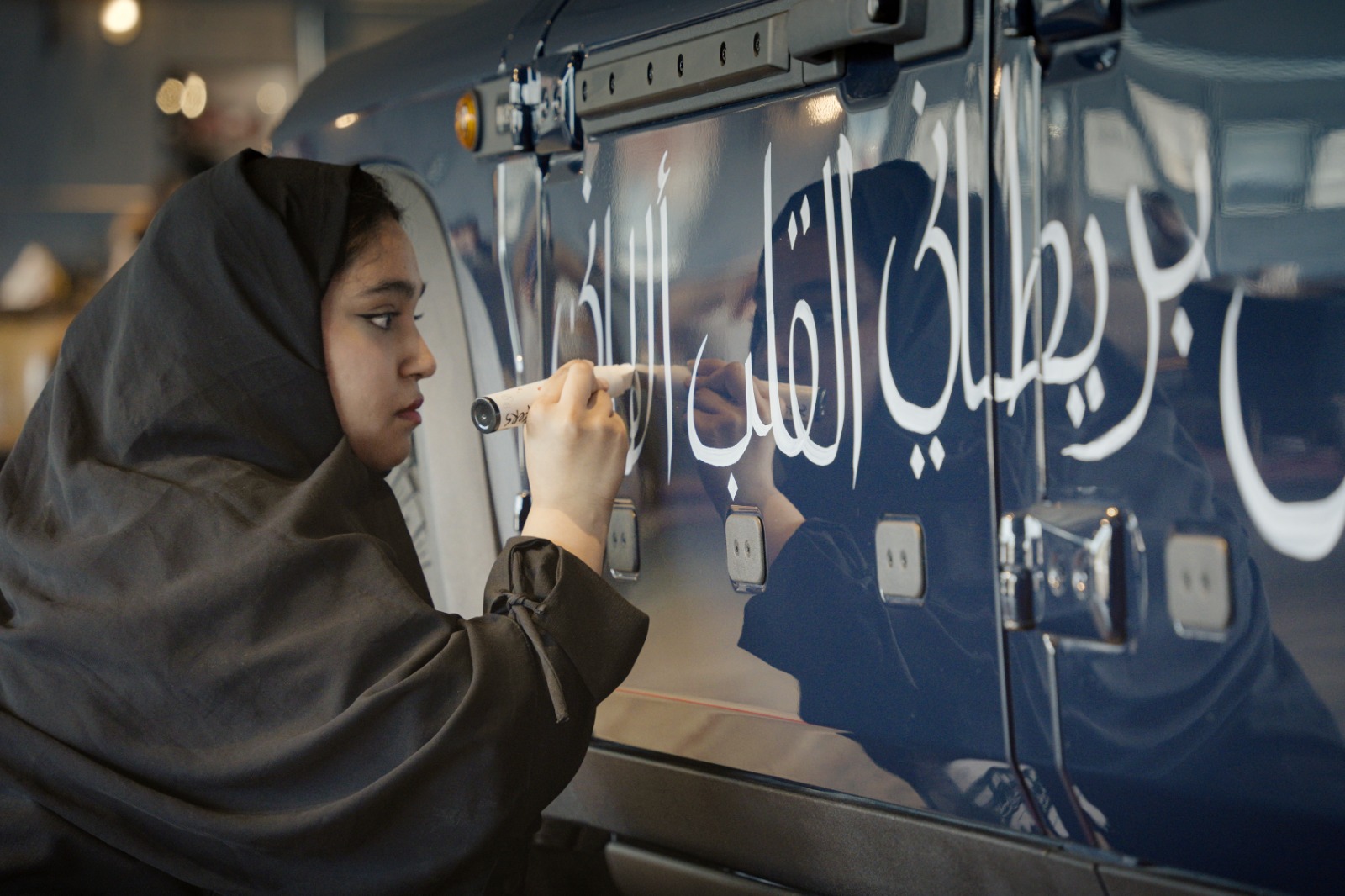 Woman writing Arabic calligraphy on the side of a blue off-road vehicle during an interactive art activity.