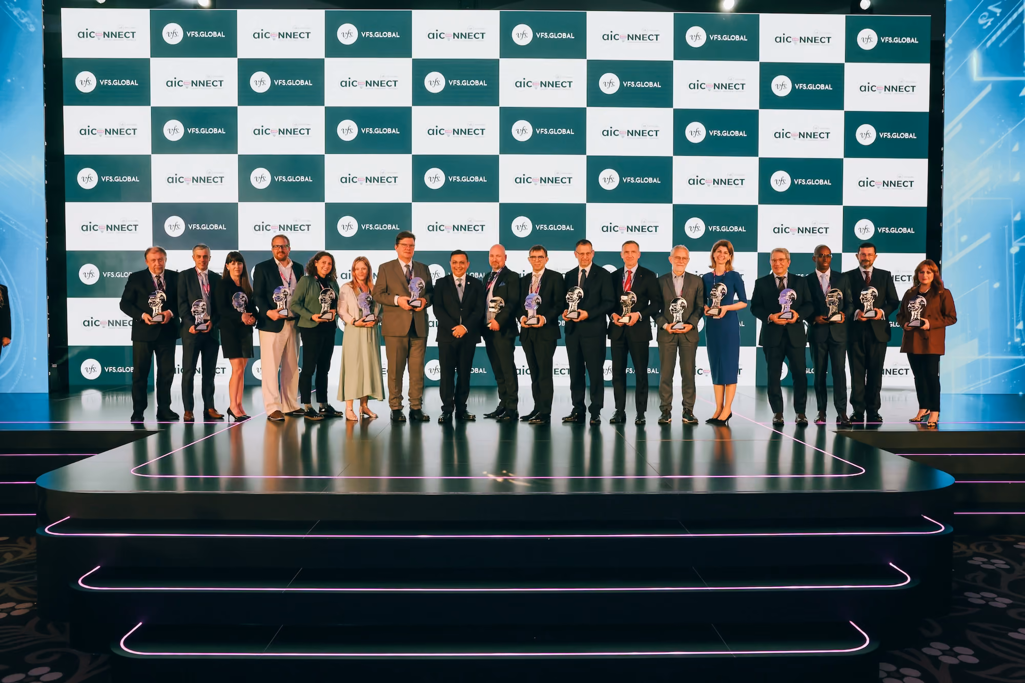 Group of award recipients holding trophies on stage at an AIC Connect and VFS Global event, with a branded backdrop behind them.