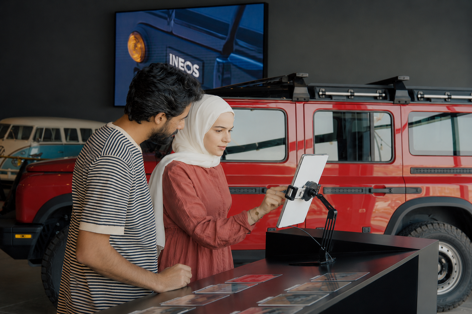 Two people exploring an INEOS vehicle display at an interactive sticker station, with a red off-road vehicle in the background.