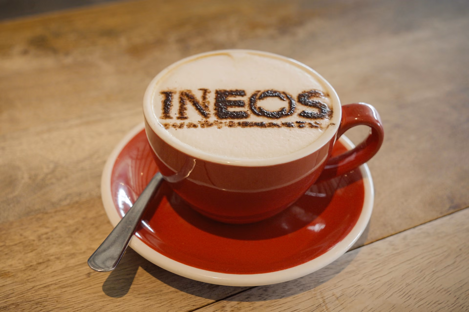 Red coffee cup with “INEOS” printed on the foam, placed on a matching saucer with a spoon on a wooden table.