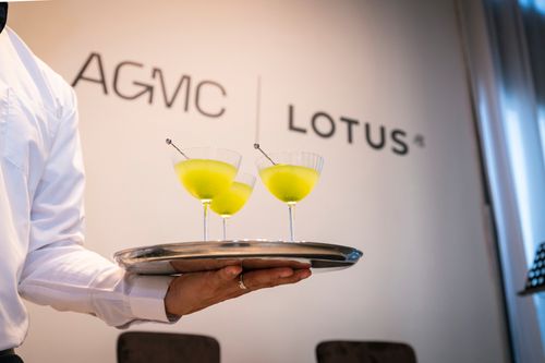 A server in a white shirt holds a silver tray with three bright yellow cocktails in ribbed glasses against an AGMC Lotus branded wall background.