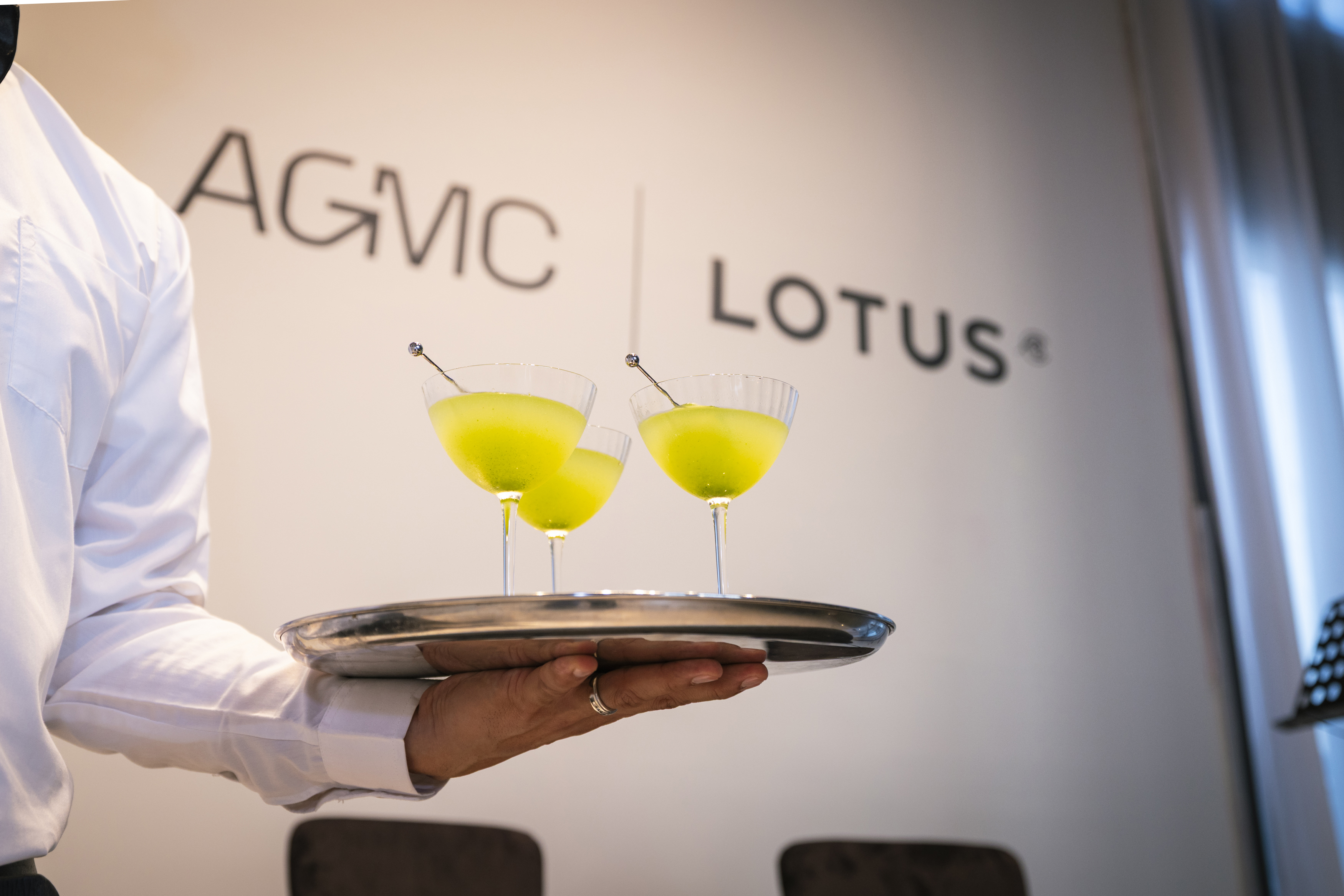 A server in a white shirt holds a silver tray with three bright yellow cocktails in ribbed glasses against an AGMC Lotus branded wall background.
