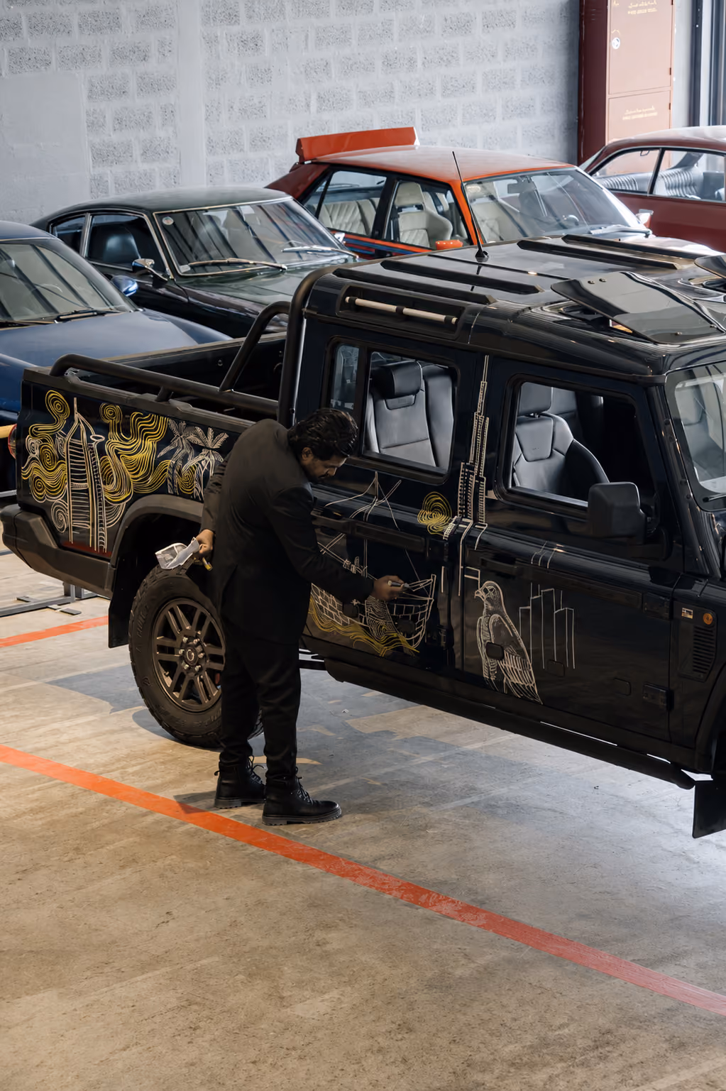 Man customizing a black off-road vehicle with intricate gold artwork, surrounded by classic cars inside an automotive workshop.